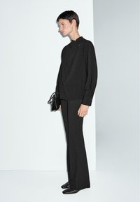Top in maglia nera con design drappeggiato, abbinato a pantaloni neri a palazzo. Estetica minimalista; include una piccola borsa a tracolla e scarpe con punta.