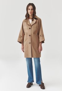 Manteau mi-long beige avec un col, de grands boutons et des poches latérales. Dispose de manches larges et d'une texture lisse. Assorti à un jean bleu et des bottes marron.