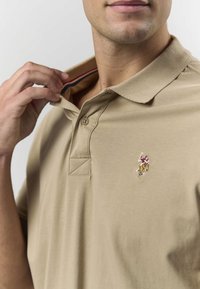 Smėlio spalvos medvilninė polo marškinėliai su trimis mygtukais, trumpomis rankovėmis ir mažu išsiuvinėtu logotipu kairėje krūtinės pusėje. Tekstūruotas apykaklė.