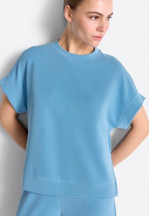 Kobieta o jasnej karnacji i związanych do tyłu włosach, ubrana w luźny, jasnoniebieski top z krótkim rękawem oraz pasujące spodnie, patrząca w prawo, z rękami za plecami.