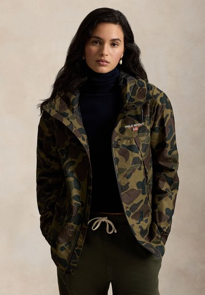 Casaco com padrão camuflado em verde, castanho e preto. Apresenta capuz, fecho de correr e logótipo bordado no peito. Textura de tecido durável.