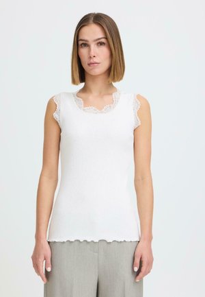 Donna con capelli lisci di media lunghezza che indossa un top bianco senza maniche con rifiniture in pizzo e pantaloni grigio chiaro, in piedi davanti a uno sfondo semplice.