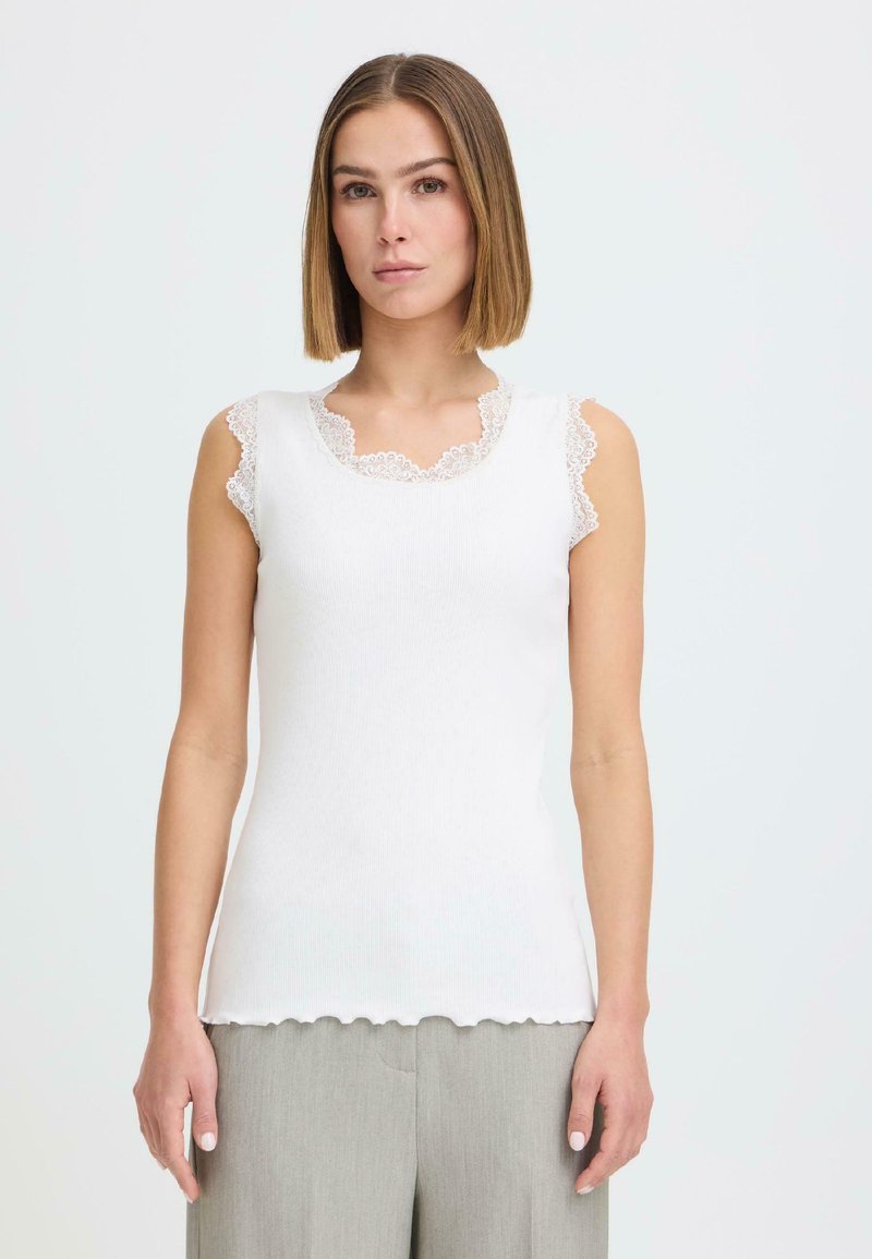 Femme aux cheveux raides mi-longs portant un haut blanc sans manches avec bordure en dentelle et un pantalon gris clair, debout devant un fond uni.