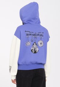 Fioletowa bluza z kapturem z białymi rękawami, z grafiką na plecach zawierającą symbole i postać. Kaptur, ściągacze na rękawach i u dołu.