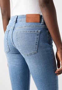 Ljusblå denimjeans med en figursydd design, som har två bakfickor och en brun läderlogga på midjebandet.