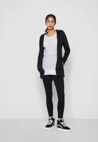Veste à capuche zippée noire avec poches, superposée à un haut en tricot blanc, associée à des leggings noirs et des baskets montantes noires avec des accents blancs.