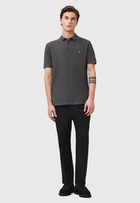 Dunkelgraues Poloshirt mit klassischem Kragen und einer Drei-Knopf-Leiste, kombiniert mit schwarzen Hosen und schwarzen Schuhen, ausgestattet mit einem dezenten Logo.