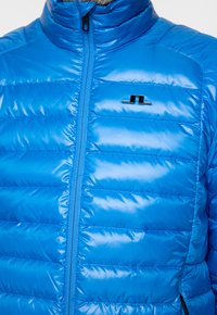 Blå puffjacka med dragkedja fram, hög krage och ett distinkt svart logotyp på bröstet. Slät, glänsande syntetisk material och quiltat mönster.