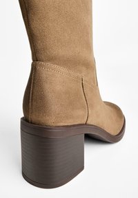 Suede brown heeled boot, featuring a chunky, textured dark brown heel and smooth rounded toe. Stitching detail on the shaft.