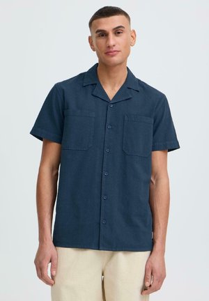 Jeune homme portant une chemise bleu marine à manches courtes avec deux poches poitrine et un pantalon beige, debout devant un fond uni clair.