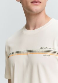 Grietos spalvos marškinėliai, pagaminti iš minkštos medvilnės, su apvaliu kaklu ir horizontalių juostelių dizainu žalią ir oranžinę, su logotipo detale kampe.