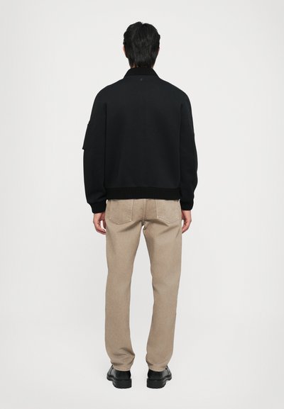 Sort sweatshirt med ribbet krave og manchetter, parret med beige bukser. Synlige bagsøm og mørke sko. Ensfarvet baggrund.