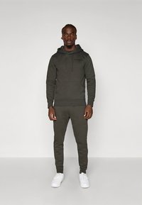 Sweatshirt verde-oliva e calças de treino combinando em tecido macio. Capuz com cordões e bolso canguru. Modelo calça ténis brancos.