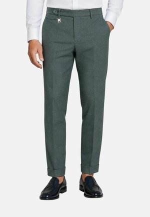 Uomo che indossa pantaloni grigi risvoltati, mocassini in pelle nera e camicia bianca a maniche lunghe, in piedi con una mano in tasca.