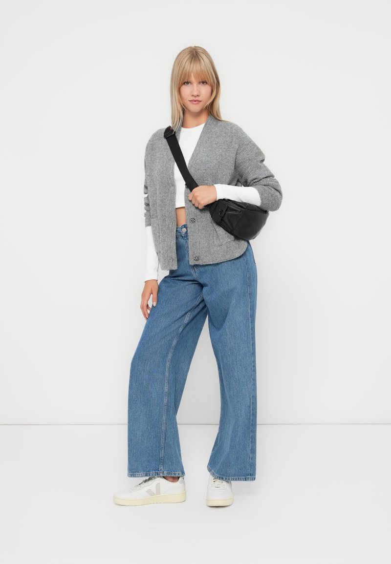 Cardigan grigio a maglia sopra una camicia a maniche lunghe bianca, abbinato a jeans blu a gamba larga e sneakers bianche. Dettaglio di una borsa a tracolla nera visibile.