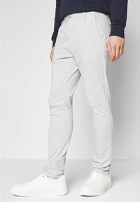 Pantaloni slim-fit gri deschis cu o textură netedă, având buzunare laterale și închidere cu nasture, asortați cu teniși albi pe un fundal neutru.