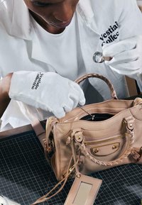 Personne portant des gants blancs et une blouse de laboratoire inspecte un sac à main en cuir beige à l’aide d’une loupe sur un tapis de découpe quadrillé.