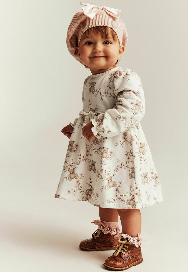Hvid kjole med blomster- og bamsemønster, lange ærmer, elastisk talje, kombineret med brune snørestøvler og en lyserød strikket baret.