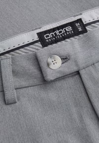 Harmaat farkut, joissa on sileä rakenne, nappikiinnitys ja etiketti, jossa lukee "ombre quintessence". Ominaisuuksiin kuuluu vyötärönauha, jossa on kontrastivärinen tikkaus.