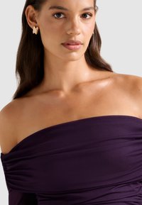 Μωβ μπλούζα εκτός ώμων με ρυθμισμένη υφή και λεία υφασμάτινη επιφάνεια. Χρυσά γλυπτικά σκουλαρίκια προσθέτου�ν μια αντίθετη λεπτομέρεια.