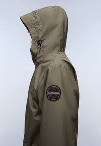 Olivengrüne Kapuzenjacke aus glattem, wasserdichtem Material. Sie verfügt über ein rundes Logo-Patch auf dem linken Ärmel und eine enge Passform.