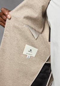 Beige Fischgrat-Anzugjacke mit taillierter Passform, Innenfutter aus leichtem Satin und weißem Etikett, das Marken- und Größeninformationen anzeigt.