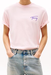Persona vestida con una camiseta de manga enrollada de color rosa claro con el logo de "Tommy" y jeans de talle alto de color azul claro, con las manos en los bolsillos.