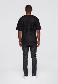 Musta lyhythihainen T-paita, jossa paljetein koristeltu siipisuunnittelu takana, yhdistetty mustiin housuihin ja valkoisiin lenkkareihin. Yksinkertainen, rento istuvuus.