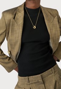 En individ i en guldfärgad blazer över en svart ribbad topp, bärande ett guldpendant halsband, poserar med händerna på höfterna. Neutral bakgrund.