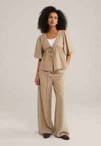 Beige, strukturiertes Oberteil mit Kordelzug in der Taille und kurzen Puffärmeln, kombiniert mit weit geschnittenen Hosen in passender Farbe und flachen, mit Nieten besetzten Slippers.