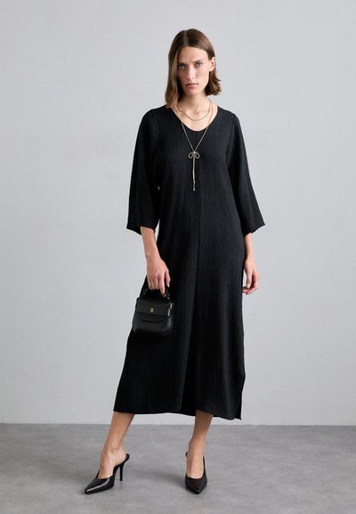 Vestido negro de corte holgado con mangas 3/4 y tela texturizada. Combinado con zapatos de tacón altos negros y un bolso pequeño.