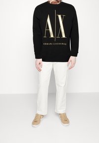 Sudadera negra con logo dorado y texto "ARMANI EXCHANGE" en el frente, combinada con pantalones blancos y zapatillas en tono beige claro.