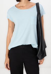 Světle modré tričko s krátkými rukávy, kulatým výstřihem a přední kapsou, spárované s černými kalhotami, drženo s texturovanou černou taškou.