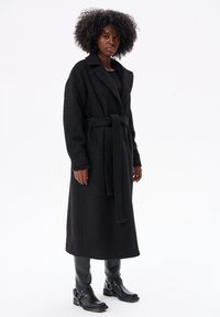 Długi czarny płaszcz wykonany z fakturowanej tkaniny z paskiem, z klapami w kształcie notcha i pełnymi rękawami; noszony na dopasowane czarne ubrania i czarne buty.