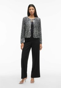 Czarna kurtka z cekinami o okrągłym dekolcie, dopasowanym kroju i otwartym przodzie, zestawiona z czarnymi spodniami o szerokich nogawkach oraz srebrnymi sandałami na obcasie.