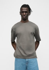 Νεαρός άνδρας με σκούρα πλεγμένα μαλλιά στέκεται με τα χέρια πίσω από την πλάτη, φορώντας απλό γκρι t-shirt και ανοιχτό μπλε τζιν παντελόνι μπροστά σε λευκό φόντο.