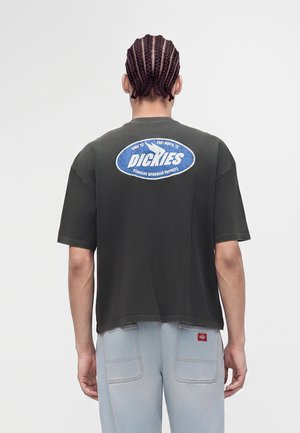 Person mit geflochtenem Haar, die ein schwarzes Dickies T-Shirt und hellblaue Jeans trägt, von hinten vor einem schlichten Hintergrund gezeigt.