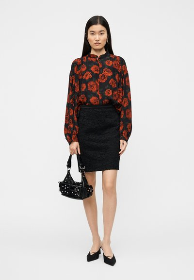Blouse fleurie noire avec motif de roses rouges, jupe crayon en dentelle noire, sac à main noir clouté, et escarpins noirs à bouts pointus. Fond uni.
