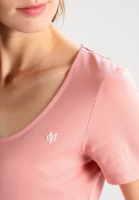 Różowa bawełniana koszulka z dekoltem w serek i krótkim rękawem, z małym białym logo wyszytym na lewej piersi. Gładka tekstura.