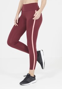 Leggings bordeaux con un alto punto vita, caratterizzati da una striscia rosa verticale lungo il lato. Abbinati a scarpe da ginnastica nere.