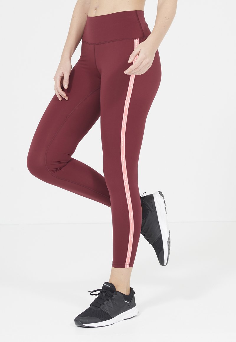 Leggings bordeaux con un alto punto vita, caratterizzati da una striscia rosa verticale lungo il lato. Abbinati a scarpe da ginnastica nere.