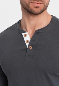 Man som bär en mörkgrå henley-tröja med vit placket och tre bruna knappar, som visar käklinjen och nacken.