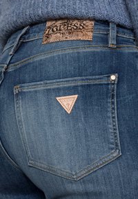 Jeans de mezclilla azul con una etiqueta marrón texturizada que presenta un diseño de estampado de serpiente en la parte trasera. Un logotipo triangular rosa está bordado en el bolsillo.