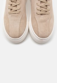 Beige mockasinskor i mocka med en slät yta, rund tå och platta snören, placerade på en vit gummisula.