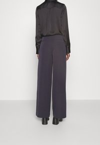 Pantalon à jambes larges en tissu bleu marine foncé à la texture lisse, doté d'une taille haute et de poches arrière discrètes. Associé à un chemisier en soie noire.