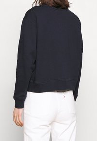Donna che indossa una felpa nera a maniche lunghe e jeans bianchi, vista di spalle su uno sfondo neutro.
