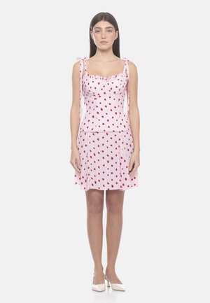 Donna in piedi che indossa un vestito rosa con pois rossi, spalline annodate e tacchi bianchi su uno sfondo bianco uniforme.