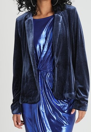 Vrouw die een donkerblauwe fluwelen blazer draagt over een glanzende, gedraaide voorkant, koningsblauwe jurk met lange mouwen.