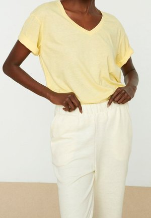 Osoba ubrana w jasnżółtą koszulkę z krótkim rękawem i dekoltem w serek oraz białe spodnie z elastycznym pasem, z rękami spoczywającymi w pobliżu talii.