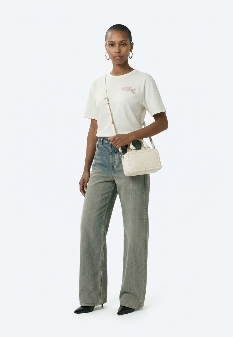 Žena u bijeloj Diesel majici, izblijedjelim širokim trapericama, crnim šiljastim potpeticama, srebrnim naušnicama u obliku obruča i s malom bijelom torbicom preko tijela.